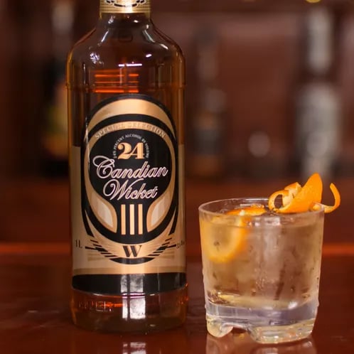 A bottle of Canadian Wicket, next to a glass with an orange peel garnish.
