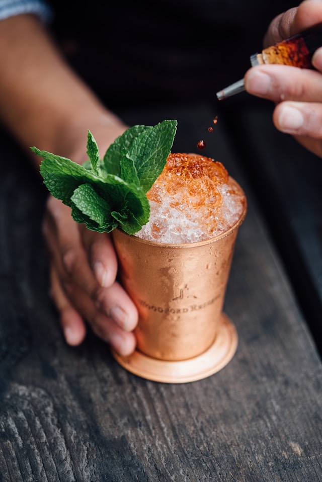 A hand drizzles syrup over crushed ice in a copper mug garnished with fresh mint on a dark wooden table.