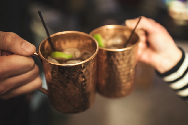 Close-up of two hands holding copper Moscow Mule mugs filled with ice and fresh lime, clinking drinks in a bar setting.