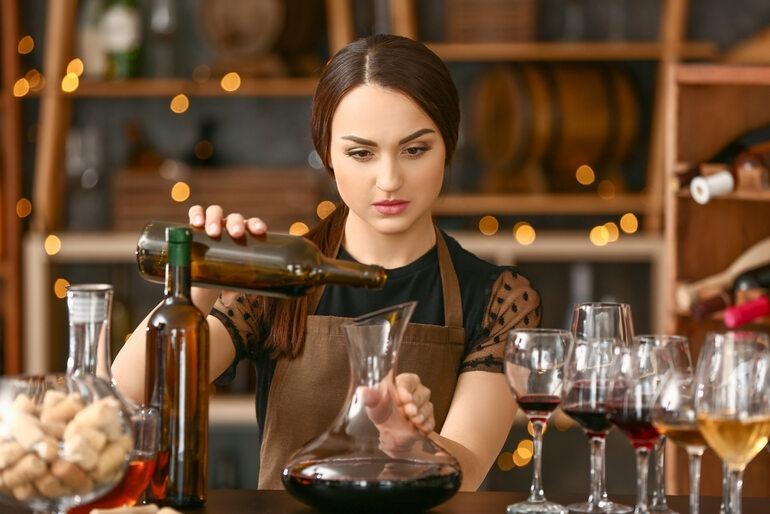 A sommelier carefully pouring red wine from a bottle into a glass decanter on a bar counter.