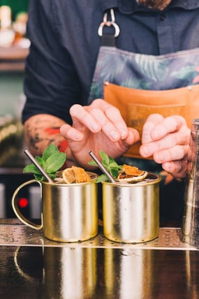 Bartender garnishing two cocktails in gold metal mugs with mint, dried citrus, and metal straws on a bar counter.